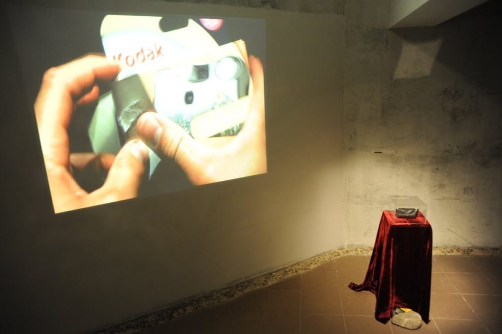 Abortus installation consisting of a projection film on a wall and the camera on a stand, with stone beside the stand